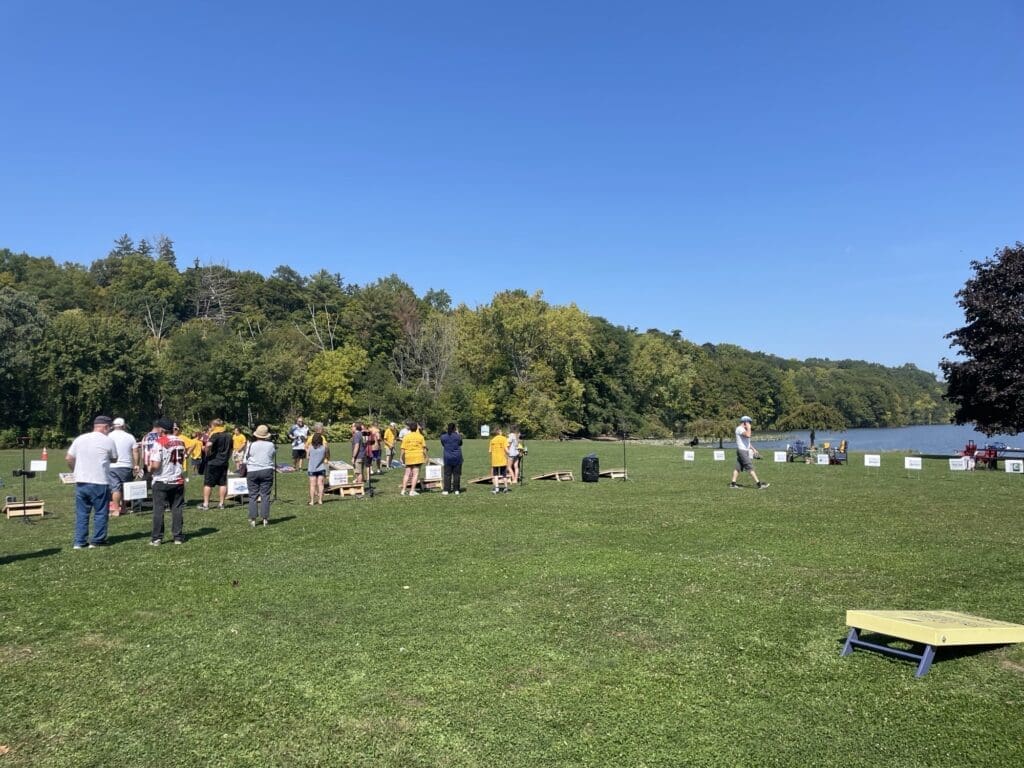 People gather outdoors on a sunny day near a wooded area and open field.